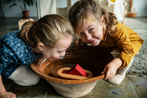 Two children playing with a wooden toy in a large bowl indoors.